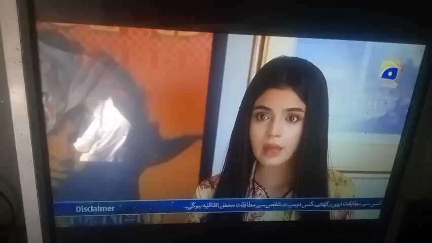The video shows a television screen displaying a scene from what appears to be a Pakistani drama. A young woman with long, dark hair is the central figure, visible from the chest up. She is wearing a light blue shalwar kameez with a matching dupatta, and her expression conveys surprise or concern. The background includes a bookshelf with various books and a mirror reflecting another individual with long, dark hair. The television screen also displays the logo of a Pakistani television channel, GEO HD, and a disclaimer in Urdu at the bottom.