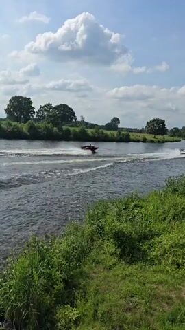 A jet ski travels down a river, leaving a wake. The water is dark and choppy. Trees and green foliage line the riverbank. The sky is blue with fluffy white clouds. The sun is shining.