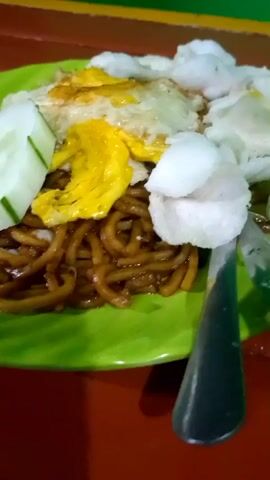 A close-up shot of a plate of food on a green plate. The food consists of noodles, a fried egg, cucumber slices, and crackers. The plate is on a red table, with a green and yellow striped background visible. The lighting is dim.