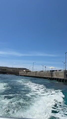 The video shows a view from a boat moving away from a pier on a sunny day. The water is turquoise and white foam is visible in the boat's wake. The pier is made of concrete blocks and has streetlights and a few people on it. There is also a small white boat docked next to the pier. The sky is blue with a few white clouds.