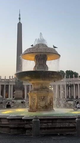 This media file captures a serene scene at St. Peter's Square in Vatican City. The central focus is a grand, multi-tiered fountain, its water cascading down in a dynamic display. Two seagulls perch atop the fountain's upper basin, adding a touch of wildlife to the urban landscape.

In the background, a towering obelisk stands tall, a significant historical monument. Behind it, the colonnades of St. Peter's Basilica create a majestic architectural backdrop. The sky is a soft gradient of pale blue and orange, suggesting either early morning or late evening light, casting a warm glow on the scene.

Several people are scattered throughout the square, some seated and others walking, enjoying the ambiance of this iconic location. The overall mood is peaceful and contemplative, with the gentle sound of the fountain and the vastness of the architecture creating a sense of awe and tranquility. The water in the lower basin of the fountain is illuminated with a soft yellow light, enhancing the magical atmosphere as dusk approaches.