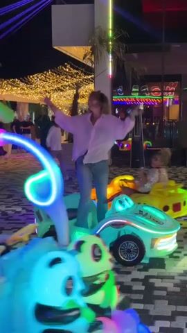 The video shows a nighttime scene at an outdoor entertainment venue. In the foreground, two young women are riding on brightly colored, illuminated amusement park rides designed to look like cartoon cars. The ride in the foreground, primarily blue and green, is in motion, and a young woman with dark hair is seated on it. To the right and slightly behind, another woman with curly reddish-brown hair, wearing a white shirt and jeans, is standing on a similar car-shaped ride, actively moving her arms and appearing to dance or cheer. Her ride is predominantly green with colorful light accents. In the background, more amusement rides are visible, and people are walking around. The environment is decorated with numerous strings of warm, twinkling lights, creating a festive atmosphere. The ground is paved with a patterned tile design. Behind the rides, there is a building with illuminated signs, some of which are neon. The overall impression is one of a lively and celebratory evening outing.