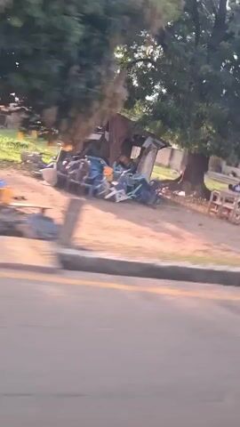 The video, apparently shot from a moving vehicle, captures a daytime scene along a road in what appears to be a tropical or subtropical location. In the initial shot, the camera is focused on the roadside, showing a pile of objects, including blue containers and metal scraps, situated near a dilapidated structure with a corrugated metal roof. As the vehicle moves forward, the camera angle shifts to reveal a man hurriedly getting onto a parked silver motorcycle, located under the structure. The sun shines brightly in the background, obscuring some details. The camera continues its motion, now capturing more of the surrounding environment. The focus moves upward to show palm trees against the sunlit sky and the rooftops of buildings.