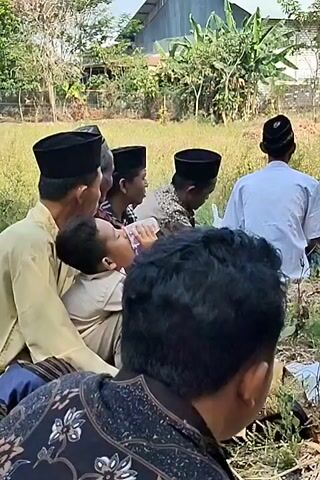 A group of men and boys are gathered outdoors in a dry, grassy area. They are all wearing traditional Islamic hats, known as peci or kopyah. Some of the men are wearing light-colored shirts and pants, while others are in darker patterned clothing. One young boy is sitting between two men and drinking from a clear plastic bottle. In the background, there are buildings with corrugated metal roofs, trees, and more people sitting under tents. The overall atmosphere appears to be casual and communal, possibly during a gathering or event.