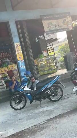 A blue and black motorcycle is parked on a concrete surface in front of a building. Two young men are near the motorcycle. One man, wearing a blue shirt, is sitting on the ground to the left of the motorcycle. He is looking towards the motorcycle. The second man, wearing a gray shirt, is sitting on the motorcycle. He is looking forward. Behind the motorcycle, there are shelves stocked with various products. Above the shelves, a sign is visible with text and images. The environment appears to be outdoors, possibly in front of a shop.