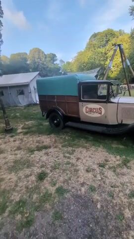 The video opens with a view of a vintage tan truck with a green tarp-covered bed and a brown wooden side panel. The truck has a "CIRCUS" logo on its side. Behind the truck, there are several tents and trees. The camera then pans to the right, showing the front of the truck with its headlights and grille. Further to the right, there's a tall tripod-like structure and some outdoor cooking equipment. The video then shifts to show a gravel path leading into a wider area with trees on either side. In the distance, a yellow and blue striped tent is visible. To the right of the path, there is a building with a covered porch and stairs. The video ends with a view of the path, the tent, and the building. The scene appears to be a circus or event set up in an outdoor, possibly wooded, area. There are no people clearly visible in the foreground, but there might be some figures in the distance near the tent.