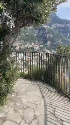 The video shows a scenic overlook of a town by the coast with mountains in the background. The perspective is from behind a black metal fence with vegetation on either side of it. The ground is paved with irregular stones. The town is densely built with white and red buildings, nestled on the hillside. The blue sea is visible along the coast. The sky is partially cloudy. The camera pans up slightly to reveal more of the town and the surrounding landscape.