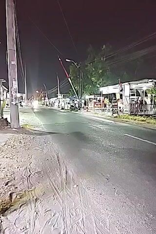 The video shows a nighttime view of a road lined with small shops and stalls on the right side. A utility pole stands prominently on the left. The road appears to be paved and has tire tracks visible on the dusty shoulder. In the distance, streetlights illuminate the road, and the headlights of a vehicle are visible. A red and white striped pole, possibly part of a barrier or gate, is positioned over the road, attached to a utility pole. A motorcycle is parked near one of the stalls. As the video progresses, a large yellow truck with a tarp-covered load drives past the camera, its headlights and taillights visible, and its movement captured with motion blur. Several people are seen walking near the stalls in the background. The overall environment suggests a rural or semi-urban area at night.