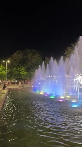 A nighttime video shows a fountain with colorful lights in the foreground. Water sprays upwards in multiple jets, illuminated by blue, purple, and green lights at the base of the fountain. Palm trees and other foliage are visible in the background, along with tents and string lights suggesting a market or event. Several people are walking in the background, and the water appears rippled, indicating some movement.