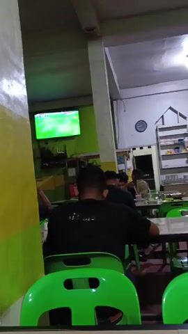 The video shows an indoor scene in what appears to be a casual dining establishment or cafe. Several people are seated at tables, some facing away from the camera. The dominant colors are green, from the chairs and a wall, and yellow, from a painted pillar. A television screen displaying a green image, possibly a sports broadcast, is mounted on the wall. There are shelves stocked with items in the background, and a clock is visible on the wall. The lighting is artificial, suggesting it is evening or indoors with no natural light source. The overall atmosphere seems relaxed, with patrons engaged in their own activities or conversations.