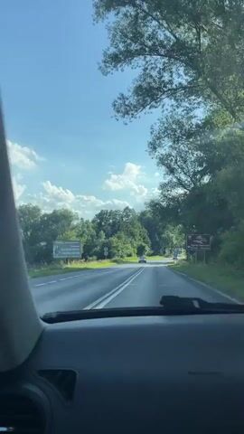 The video shows a first-person view from inside a car driving on a road surrounded by trees. The sky is bright blue with some fluffy white clouds, suggesting it is daytime with good weather. The road appears to be a two-lane highway, and there are other cars on the road ahead. 

On the right side of the road, there are two signs. One sign, with a blue background and white text, shows a speed limit of 50 km/h and a parking symbol. Another sign, with a white background and black text, indicates "Ośrodek Japoński" with an arrow pointing right and a distance of 12 km to "Słoneczna". On the left side of the road, another sign with a green background and white text reads "GALERIATwierdzenia" followed by some Polish text that is difficult to read, and below it "CYNDRON DOMOWE".

The overall impression is a pleasant drive through a rural or suburban area in Poland. The mood seems relaxed and peaceful.
