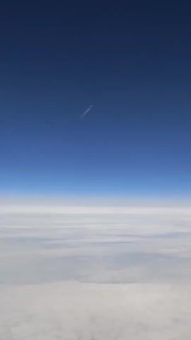 The video starts with a shot of a fluffy expanse of white clouds, viewed from above, with a clear blue sky overhead. A jet plane is visible in the distance, leaving a white contrail. The camera zooms in on the plane as it flies diagonally across the sky, its contrail stretching behind it. The view then pans up, focusing on the blue sky as the plane continues its journey, eventually disappearing from sight.