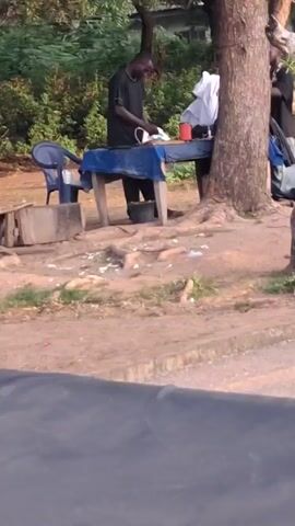 The video shows a man standing behind a makeshift ironing board set up outdoors, presumably ironing clothes. The ironing board is a wooden table covered with a blue cloth. Beside him, a bucket sits on the ground. To the left is a blue chair, and a small wooden stool. A large tree stands next to the table. Another man joins the first man behind the table, and a young woman walks past them. The scene is situated on a dirt patch next to a road with greenery visible in the background. The video is shot from a low angle, possibly from inside a vehicle.