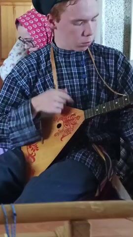 The video shows three people playing balalaikas. The first person, a young man, is wearing a blue and black plaid shirt and dark pants. He has reddish-brown hair. Behind him to the left is a person wearing a red headscarf with a white flower pattern and glasses. To the right of the young man is another person, also playing a balalaika. The environment appears to be inside, possibly a home or venue decorated in a traditional style. There are wooden structures and decorations visible in the background. The main action is the playing of balalaikas, and the people seem to be engaged in a musical performance.