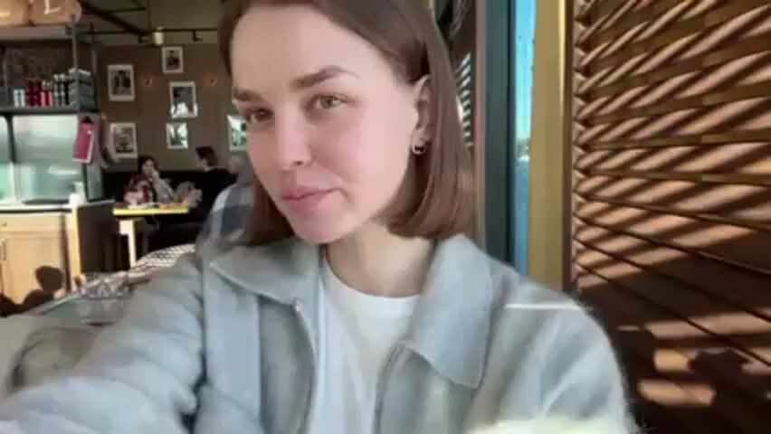 A young woman with short brown hair wearing a white t-shirt and a grey zip-up sweater sits at a table in a restaurant, holding up a piece of flatbread with toppings and smiling. Sunlight streams in through a slatted window behind her, casting shadows. Other patrons are visible in the background, sitting at tables and interacting. A shelf stocked with various items is also visible in the background.