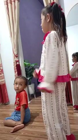 A young child in an orange shirt and blue shorts sits on the floor looking to the left, with their back mostly to the camera. Another young child, taller and wearing a matching pajama set with a white base and pink trim and a pattern of small flowers and vertical lines, stands facing away from the camera, looking to the left. The standing child is in the process of dancing or moving their arms. The background shows a wooden floor, a curtain on the left, and a large mirror on the right reflecting another child in a similar outfit. A potted plant is visible in the background.