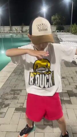 The video captures a young boy dancing by a swimming pool at night. He is wearing a white t-shirt with a "B.Duck" graphic, red shorts, a baseball cap, and sneakers. The boy is performing dance moves with his arms and body.

The scene is set outdoors next to a swimming pool illuminated by lights. In the background, there are lounge chairs and some vegetation. The time of day is clearly night, indicated by the darkness and the artificial lighting. The weather appears to be clear, as there are no visible signs of rain or wind.

The boy's stance and energetic movements suggest he is enjoying himself and possibly performing a choreographed dance. The overall atmosphere is casual and playful, with the boy being the central focus of the video. The visible text on the boy's t-shirt reads "B.DUCK" in large, bold letters. His cap has "NYC DOWNTOWN" printed on it. The shorts also have text that appears to read "CHILLOUT" vertically along the side. The location context of Giachrypsh, Abkhazia, places the scene in a resort or recreational area.