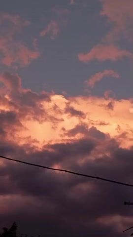 A time lapse video shows a sunset with pink and orange clouds against a dark purple and blue sky. A power line is visible across the middle of the frame. On the right side of the frame, a streetlamp and an antenna appear. At the bottom of the frame, the tops of trees are visible.