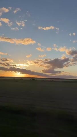 The video shows a scenic sunset over a large field. The sky is blue with scattered white clouds, some tinged with golden hues from the setting sun. The sun is low on the horizon, partially obscured by a layer of clouds, casting a warm, golden light across the sky and landscape. Below, a vast green field stretches out to the horizon, bordered by a dark line of trees or vegetation in the distance. There are no people visible in the shot. The camera appears to be stationary, capturing the serene beauty of the sunset over the field.