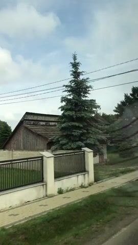 The video, shot from a moving vehicle, displays a rural or semi-rural landscape. A paved path runs alongside a green wall or fence, beyond which are grassy areas and various trees. Several houses with different roof styles, including red and dark gray, are visible. Power lines and utility poles span the scene. A yellow front-end loader is parked near one of the houses. A park bench is briefly visible next to the green wall. The sky is partly cloudy.