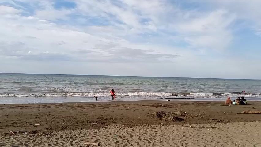 A wide shot of a beach with a person and a child standing in the shallow water on the left, and a group of people sitting on the sand on the right. The ocean is calm with small waves. The sky is blue with scattered white clouds. The sand is dark brown and appears to be wet near the water's edge. There is some debris scattered on the sand, including driftwood.