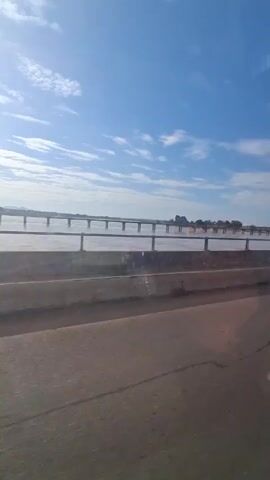 The video shows a view of a bridge over water from the inside of a moving vehicle. The sky is blue with some white clouds. The water is murky. A concrete barrier runs along the side of the road. The road surface is asphalt. The frame of the car is visible on the edges of the shot. Sounds of children singing can be heard.