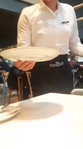 A server wearing a white shirt and a "The Glenlivet" apron places a round, dark-brown dessert on a white plate onto a white table. A wooden condiment caddy with an oil bottle and salt and pepper shakers sits next to the plate. Patrons are seated in the background.