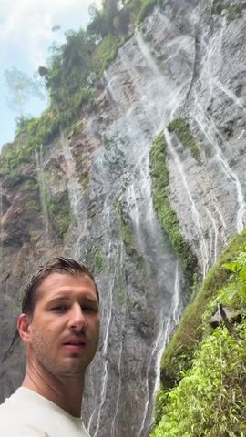 A man stands in front of a tall waterfall, the water cascading down a rocky cliff face. Lush green vegetation surrounds the waterfall and climbs the cliff. The man is looking directly at the camera with a neutral expression. He is wearing a white t-shirt. The video is a close-up shot focusing on the man and the waterfall behind him.
