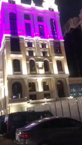 The video shows a tall, light-colored building at night, brightly lit with both white and pink lights. The building has a classic architectural style with arched windows and decorative molding. Several cars are parked in front of the building. The camera is angled upwards, focusing on the building's facade. The surrounding environment is dark, with a glimpse of another tall, dark building in the background. There is no visible activity or people in the shot.