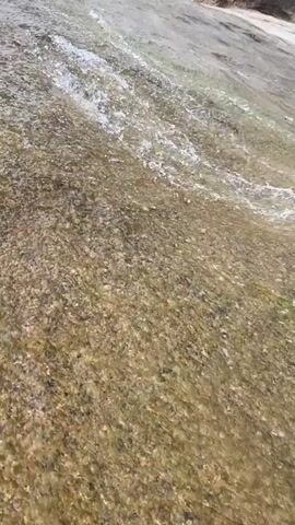 The video shows a close-up of water flowing over a rocky surface. The water is clear and shallow, allowing the viewer to see the rocks beneath. There is foam visible on the water surface. The camera angle is slightly tilted. A person's leg and foot wearing a dark colored shoe are briefly visible at the top right corner of the frame.