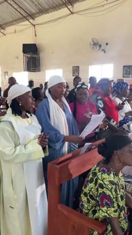 Inside a church, a choir sings together. The choir consists of men and women, many of whom are holding sheets of music or songbooks. Several women wear headscarves, and some are dressed in African print clothing. A conga player accompanies the choir, while a conductor leads them. The church interior features light-colored walls, windows with natural light, and visible wiring along the ceiling. A few framed pictures hang on the wall. The overall scene is a religious gathering with music and singing at its core.