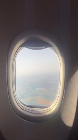 The video shows an airplane window with a view of the landscape below. The window is oval-shaped and framed by a metallic rim. The view outside is of green fields and some scattered trees, suggesting a rural or suburban area. The sky is hazy and light blue, with the sun casting a bright glow on the right side of the window. The interior of the airplane cabin is dark and appears to be out of focus, with a hint of a seat or fabric on the right side. The camera is positioned inside the plane, looking out through the window. The video is static, with no apparent movement of the plane. The lighting suggests it is daytime, possibly late afternoon or early morning given the angle of the sun.