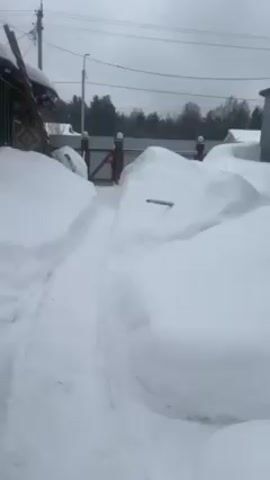 The video begins with a view of a snow-covered path leading toward a gated fence. The path has been cleared, leaving high snowdrifts on either side. The camera then pans to the left, revealing a wooden structure with a snow-covered roof. The camera then comes to rest on a woman wearing a large, hooded winter coat. Snowflakes are visible in the air. She looks directly at the camera and smiles. The background includes trees and houses, all covered in snow. The sky is overcast.