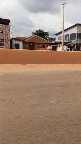 The video shows a roadside scene in what appears to be a developing area. The road is dusty and lined with concrete barriers. In the background, there are various buildings, including a two-story structure with large windows, a smaller building with a sign, and several residential-type houses. There are also utility poles and power lines visible. 

In the latter part of the video, a motorcycle with two people on it drives across the road, kicking up dust as it moves.