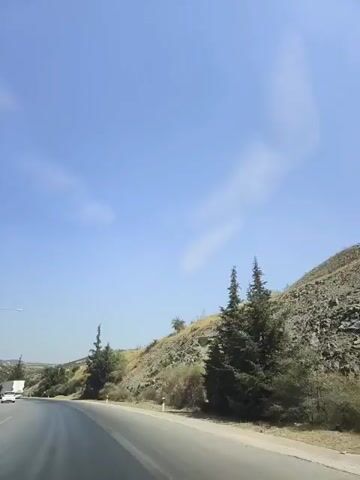 The video shows a view from a moving vehicle on a road. The road curves to the left and is bordered by a rocky, dry hillside on the right. There are scattered trees and shrubs on the hillside, including several cypress trees. The sky is clear and blue with a few wispy clouds. In the distance on the left side of the road, there is a white truck or van. The road surface appears to be asphalt and shows signs of wear. The overall environment appears to be rural or semi-rural.