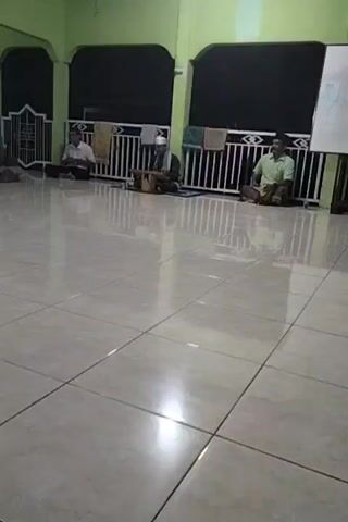 The video depicts three men sitting on the floor in a room with a tiled floor and green walls. The men are spaced apart from each other. One man in the middle is playing a stringed instrument. Towels are hanging on a white railing behind them. A whiteboard stands on the right side of the frame. The room appears to be a mosque or similar gathering space.