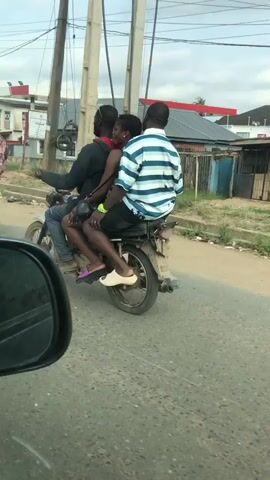 The video shows three people on a motorcycle. The person in front is driving. A woman is in the middle, and another person is behind her. They are on a paved road with buildings and a gas station in the background.