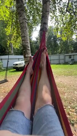 A first-person perspective video shows a person lying in a hammock. The hammock is made of colorful fabric, with stripes of red, yellow, green, and blue. The person is wearing denim shorts. Their bare feet are visible at the top of the frame, and their legs extend down into the hammock. The hammock is suspended between two birch trees. Sunlight filters through the leaves of the trees, dappling the scene with light and shadow. In the background, a white SUV is parked on a grassy area, and a white fence runs along the edge of the property. The overall environment appears to be a backyard or outdoor recreational area on a sunny day.