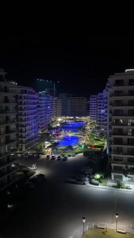 This aerial, nighttime shot captures a modern resort complex in Gastria, Northern Cyprus. The scene is dominated by sleek, multi-story white buildings with numerous balconies, illuminated by purple and white lights that accentuate their architectural design. Between the buildings, a vibrant pool area glows with bright blue lights, surrounded by greenery and pathways. Cars are parked in designated lots, also illuminated by spotlights. The overall atmosphere is one of modern luxury and nighttime entertainment. The sky is dark, indicating it is late at night. The resort appears to be a popular destination, judging by the number of lights and the well-maintained grounds.