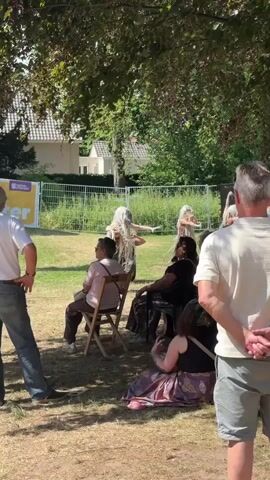 A video captures a performance by individuals dressed in elaborate, white, flowing costumes and wigs, with some wearing headpieces resembling animal skulls. They appear to be dancing or performing a ritualistic movement outdoors. Several audience members are seated on chairs and on the ground, facing the performers. The setting is a grassy area with trees providing shade. In the background, there is a fence, some vegetation, and buildings. The lighting suggests it is daytime, likely sunny. The camera perspective is from behind the audience, looking towards the performers.