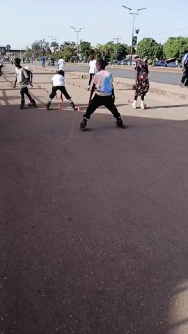 A medium shot shows four people roller skating on a paved road. A row of orange cones is in the background. There are trees, lamp posts, cars and other people in the distance. One skater is bent over. The other three are upright. The person closest to the camera skates a few steps forward. All are wearing skates and casual clothing.