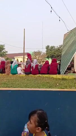 The video shows a group of people sitting on a raised ledge. In the foreground, a young child with dark hair pulled into two pigtails, wearing a yellow shirt, looks towards the left side of the frame. Behind the child is a blue surface, possibly a wall or barrier. Beyond the ledge, the group of people consists of individuals wearing various colors of clothing, many of whom are wearing hijabs. There are trees and buildings in the background, suggesting an outdoor setting. A street light is visible on the left side of the frame. String lights are hanging from the top right corner of the frame.