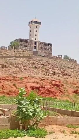 The video shows a partially constructed building on a rocky hill. The building appears to be a tower with multiple stories. In the foreground, there are some plants and a dirt path. The sky is clear and the sun is shining. The video is shot from a slightly elevated position, looking up at the building. The camera is panning slowly to the right.
