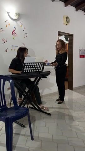 The video shows a group of women gathered indoors to sing, likely in a rehearsal setting.  

A woman is playing a keyboard while a woman in black, holding a tray, walks past. The camera moves to show a group of women seated and holding sheet music. The background includes a pink curtain with a musical note design. The room has white walls and tiled floors. There are musical notes on the wall next to the keyboard. The women appear to be actively singing, looking at their sheet music, their mouths are visibly moving. One woman has gray hair.