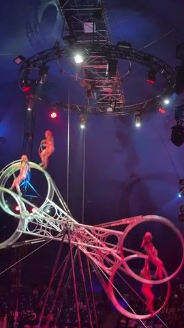 The video shows a circus performance taking place inside a large tent. The primary focus is on a complex metal apparatus with several large rotating rings. Several performers, dressed in red and gold costumes, are actively engaged in acrobatic feats on and around these rings. Some performers are seen running inside the rings as they spin, while others are balancing precariously on the edges. In the background, there is a circular lighting rig suspended from the ceiling, illuminating the performance area with red and white lights. The audience is visible in the lower portion of the frame, seated in rows of chairs. The overall atmosphere is one of excitement and athletic prowess.
