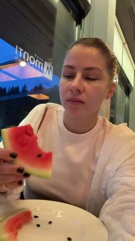 A young Caucasian woman is shown in a close-up shot, sitting indoors with a plate of watermelon in front of her. She is wearing a white t-shirt and a white open shirt. Her hair is pulled back. She is holding a slice of watermelon to her mouth and appears to be eating it. There are watermelon seeds on the plate. In the background, through a window, a dark sky with trees and a sign with the word "Monti" is visible. The lighting suggests it is evening.