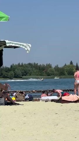 This is a medium shot video taken on a sunny day at a beach. The foreground shows a sandy beach with several people sunbathing and relaxing on towels and lounge chairs. Some people are sitting on a wooden pier that extends into a body of water. In the background, the water is blue and there are trees and greenery along the shore. There are several watercraft on the lake including what appears to be a yellow inflatable tube and a jet ski. The overall atmosphere appears to be one of leisure and recreation.