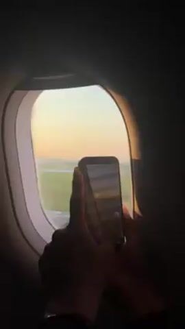 From the perspective of an airline passenger, a person holds a smartphone up to an airplane window, recording a video of the scene outside. The window frames a view of the runway at dawn or dusk, with a golden sun illuminating the scene. The airplane appears to be moving.