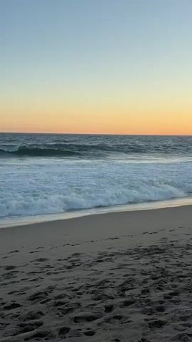A wide shot shows a sandy beach in the foreground with footprints visible. Gentle waves with white foam are rolling in on the ocean. The sky above is clear with a gradient of pale blue at the top transitioning to a warm orange hue near the horizon, suggesting either sunrise or sunset. The water is a deep blue.