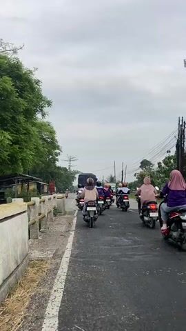 The video shows a group of people riding motorcycles on a paved road. The sky is overcast, and there are trees and some buildings visible along the side of the road. The motorcycles are traveling in the same direction, and the riders are mostly wearing casual clothing, some with helmets. The camera is positioned behind the motorcycles, following their movement. There is a large truck in front of the motorcycles, also moving in the same direction. The road appears to be a rural or suburban area, with power lines visible overhead. The overall impression is of a mundane commute or journey.